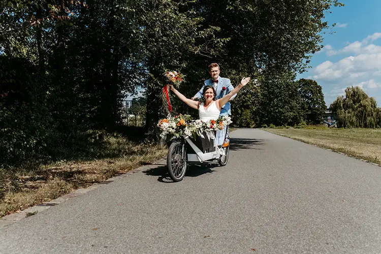 Ein fröhliches Paar fährt mit einem blumengeschmückten Lastenfahrrad eine sonnige Straße in der Nähe von Schloss Albrechtsberg in Dresden entlang. Die Braut im weißen Kleid hält einen Blumenstrauß in der Hand und streckt freudig die Arme in die Höhe. Der Bräutigam im Anzug steht lächelnd hinter ihr, während grüne Bäume die Straße säumen.