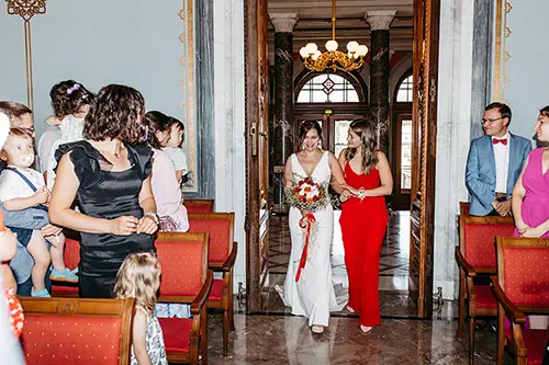 Im eleganten Saal von Schloss Albrechtsberg Dresden schreitet eine Braut im weißen Kleid mit ihrer Brautjungfer in Rot anmutig zum Altar. Gäste auf roten Stühlen halten den Moment fest, während Melanie Kunert Fotografie diese wunderschöne Hochzeit unter dem schimmernden Kronleuchter verewigt.