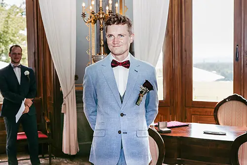 Eine Person in einem hellblauen Anzug mit roter Fliege steht lächelnd auf dem Hochzeitsgelände Schloss Albrechtsberg in Dresden, umgeben von kunstvollem Dekor. Im Hintergrund ist eine weitere Person in einem dunklen Anzug zu sehen. Licht strömt durch große Fenster. Eingefangen von Melanie Kunert Fotografie, wirkt die Szene elegant und ruhig.