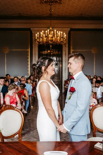 Bei ihrer HOCHZEIT in Schloss Albrechtsberg gibt sich ein Paar in einem eleganten Raum das Ja-Wort. Der Bräutigam, in hellblauem Anzug und roter Fliege, hält eine Rose in der Hand. Die Braut glänzt in ihrem weißen Kleid mit aufwendigen Haardetails, während die Gäste, darunter ein Kind, aufmerksam im Hintergrund zuschauen.