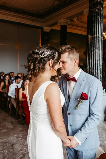 Bei ihrer HOCHZEIT in Schloss Albrechtsberg gibt sich ein Paar in einem eleganten Raum das Ja-Wort. Der Bräutigam, in hellblauem Anzug und roter Fliege, hält eine Rose in der Hand. Die Braut glänzt in ihrem weißen Kleid mit aufwendigen Haardetails, während die Gäste, darunter ein Kind, aufmerksam im Hintergrund zuschauen.
