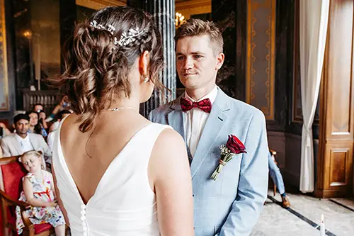 Bei ihrer HOCHZEIT in Schloss Albrechtsberg gibt sich ein Paar in einem eleganten Raum das Ja-Wort. Der Bräutigam, in hellblauem Anzug und roter Fliege, hält eine Rose in der Hand. Die Braut glänzt in ihrem weißen Kleid mit aufwendigen Haardetails, während die Gäste, darunter ein Kind, aufmerksam im Hintergrund zuschauen.