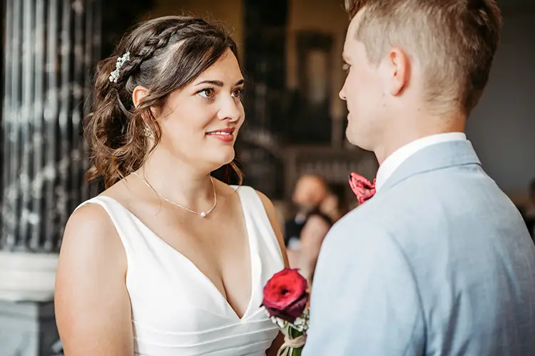 Braut und Bräutigam stehen sich im eleganten Ambiente von Schloss Albrechtsberg mit herzlichem Lächeln gegenüber. Die Braut trägt ein weißes Kleid, ihr Haar ist elegant frisiert, und sie hält eine rote Rose in der Hand. Der Bräutigam trägt einen hellgrauen Anzug. Die beiden stehen in diesem Dresdner Ambiente und lassen eine fröhliche Hochzeitsfeier erwarten.