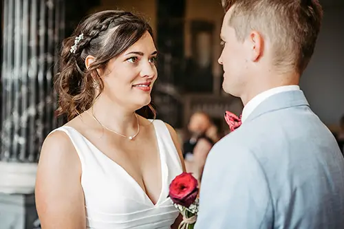 Braut und Bräutigam stehen sich im eleganten Ambiente von Schloss Albrechtsberg mit herzlichem Lächeln gegenüber. Die Braut trägt ein weißes Kleid, ihr Haar ist elegant frisiert, und sie hält eine rote Rose in der Hand. Der Bräutigam trägt einen hellgrauen Anzug. Die beiden stehen in diesem Dresdner Ambiente und lassen eine fröhliche Hochzeitsfeier erwarten.