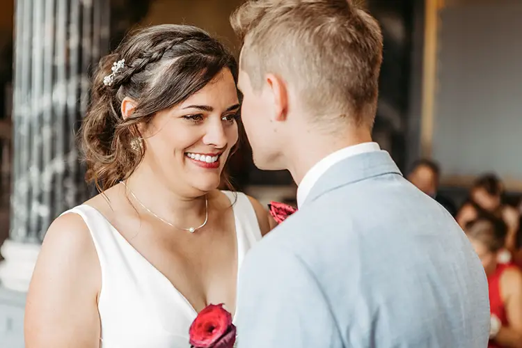 Braut und Bräutigam stehen sich im eleganten Ambiente von Schloss Albrechtsberg mit herzlichem Lächeln gegenüber. Die Braut trägt ein weißes Kleid, ihr Haar ist elegant frisiert, und sie hält eine rote Rose in der Hand. Der Bräutigam trägt einen hellgrauen Anzug. Die beiden stehen in diesem Dresdner Ambiente und lassen eine fröhliche Hochzeitsfeier erwarten.