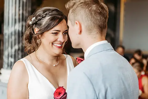 Braut und Bräutigam stehen sich im eleganten Ambiente von Schloss Albrechtsberg mit herzlichem Lächeln gegenüber. Die Braut trägt ein weißes Kleid, ihr Haar ist elegant frisiert, und sie hält eine rote Rose in der Hand. Der Bräutigam trägt einen hellgrauen Anzug. Die beiden stehen in diesem Dresdner Ambiente und lassen eine fröhliche Hochzeitsfeier erwarten.