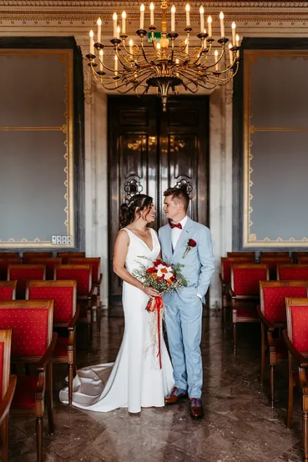 Braut und Bräutigam stehen in einem eleganten Raum im Schloss Albrechtsberg Dresden, geschmückt mit kunstvollen Kronleuchtern und roten Stühlen. Die Braut hält einen leuchtenden Strauß roter und weißer Blumen in den Händen, während sie sich liebevoll anschauen – wunderschön eingefangen von Melanie Kunert Fotografie.