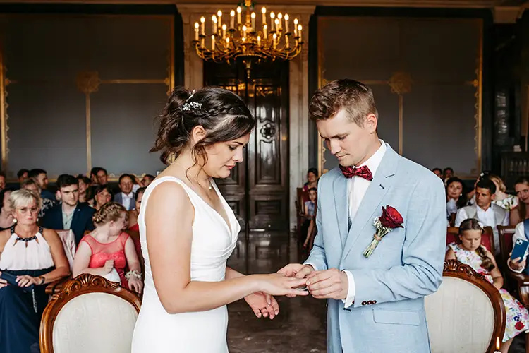 Während ihrer Hochzeit auf Schloss Albrechtsberg in Dresden tauscht ein Paar in einem eleganten, von Kerzenlicht erleuchteten Raum die Ringe. Das weiße Kleid der Braut und der hellblaue Anzug des Bräutigams mit roter Fliege fangen den Moment wunderschön ein, dokumentiert von Hochzeitsfotografin Dresden Melanie Kunert Fotografie.