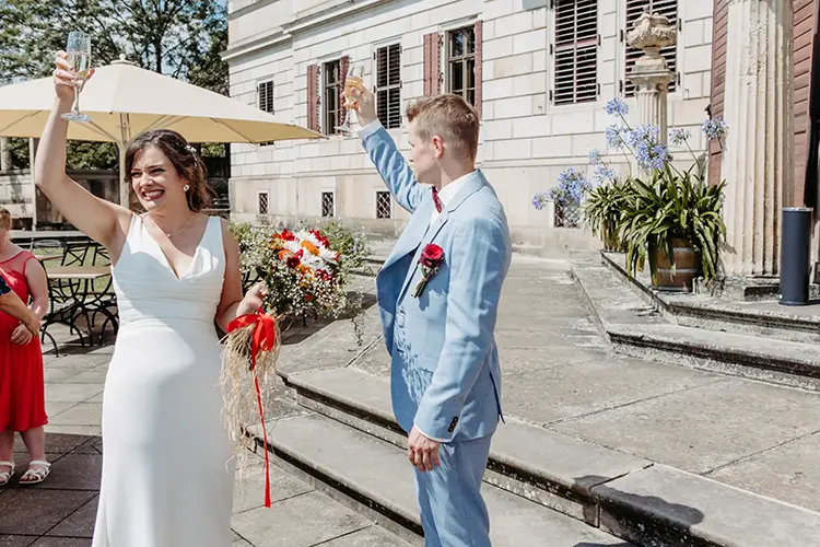 Inmitten der Eleganz von Schloss Albrechtsberg lächelt eine Braut im weißen Kleid, hält einen Blumenstrauß und erhebt ein Champagnerglas. Neben ihr steht der Bräutigam im hellblauen Anzug. Melanie Kunert Fotografie fängt diesen freudigen Moment vor der Kulisse von Blüten und Sonnenschirmen ein und feiert die Liebe in Dresden.