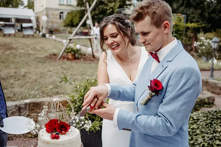 Ein Paar schneidet seine weiße Hochzeitstorte an, die mit roten Rosen und Schleierkraut verziert ist. Der Bräutigam trägt einen hellblauen Anzug, während die Braut in ihrem weißen Kleid glänzt. Der „Mr. & Mrs.“-Aufsatz rundet diesen zauberhaften Hochzeitsmoment ab.