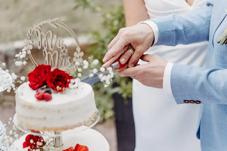 Ein Paar schneidet seine weiße Hochzeitstorte an, die mit roten Rosen und Schleierkraut verziert ist. Der Bräutigam trägt einen hellblauen Anzug, während die Braut in ihrem weißen Kleid glänzt. Der „Mr. & Mrs.“-Aufsatz rundet diesen zauberhaften Hochzeitsmoment ab.