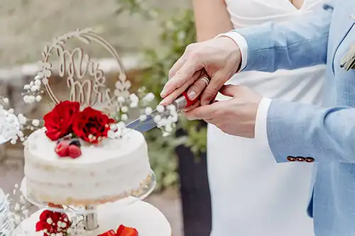 Ein Paar schneidet seine weiße Hochzeitstorte an, die mit roten Rosen und Schleierkraut verziert ist. Der Bräutigam trägt einen hellblauen Anzug, während die Braut in ihrem weißen Kleid glänzt. Der „Mr. & Mrs.“-Aufsatz rundet diesen zauberhaften Hochzeitsmoment ab.