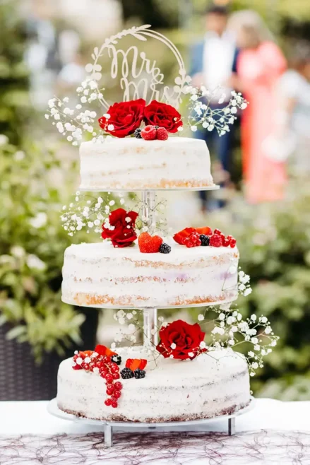 Eine dreistöckige Hochzeitstorte mit minimalistischem Design, verziert mit roten Rosen, Schleierkraut, Himbeeren, Brombeeren und roten Johannisbeeren. Ein „Mr & Mrs“-Aufsatz krönt die Torte vor einem Hintergrund aus verschwommenem Grün.