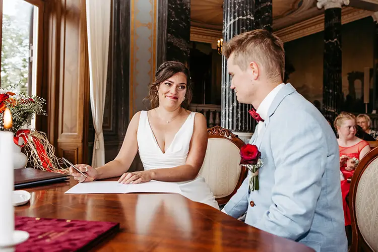 Bei der Hochzeit auf Schloss Albrechtsberg in Dresden sitzen eine Braut im weißen Kleid und ein Bräutigam im hellblauen Anzug am Tisch. Anmutig unterschreibt sie ein Dokument, während im Hintergrund die Gäste sitzen.