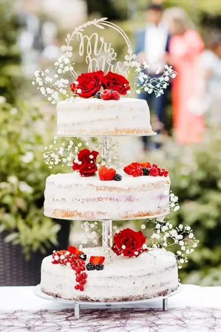 Eine dreistöckige Hochzeitstorte mit minimalistischem Design, verziert mit roten Rosen, Schleierkraut, Himbeeren, Brombeeren und roten Johannisbeeren. Ein „Mr & Mrs“-Aufsatz krönt die Torte vor einem Hintergrund aus verschwommenem Grün.