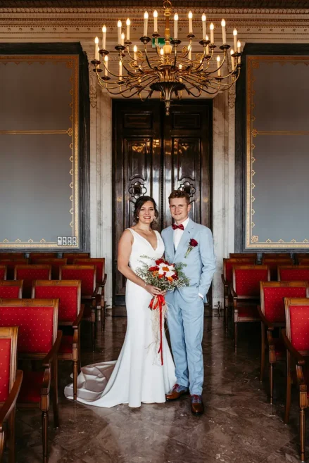 Braut und Bräutigam stehen in einem eleganten Raum im Schloss Albrechtsberg Dresden, geschmückt mit kunstvollen Kronleuchtern und roten Stühlen. Die Braut hält einen leuchtenden Strauß roter und weißer Blumen in den Händen, während sie sich liebevoll anschauen – wunderschön eingefangen von Melanie Kunert Fotografie.