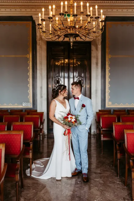 Braut und Bräutigam stehen in einem eleganten Raum im Schloss Albrechtsberg Dresden, geschmückt mit kunstvollen Kronleuchtern und roten Stühlen. Die Braut hält einen leuchtenden Strauß roter und weißer Blumen in den Händen, während sie sich liebevoll anschauen – wunderschön eingefangen von Melanie Kunert Fotografie.