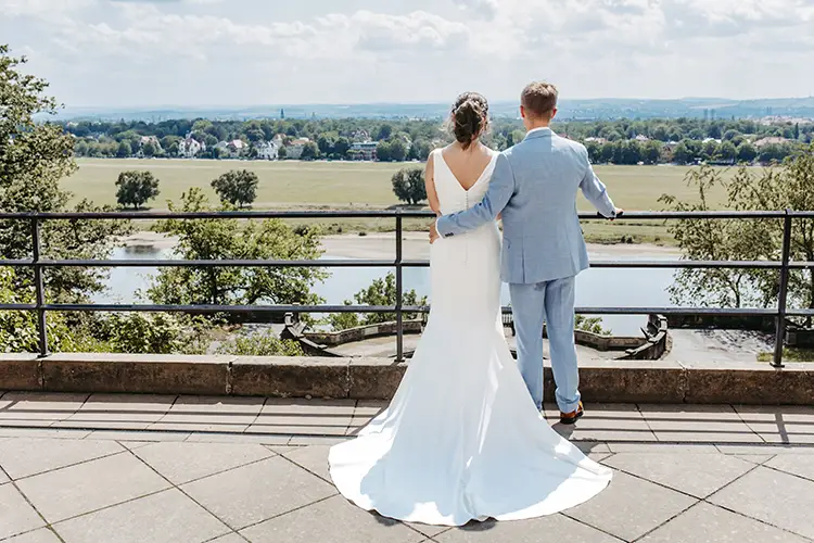 Braut und Bräutigam stehen Seite an Seite und sind fasziniert vom malerischen Blick auf den Fluss vor Schloss Albrechtsberg in Dresden. Die Braut glänzt in ihrem weißen Kleid, während der Bräutigam sie in einem hellblauen Anzug ergänzt. Wunderschön eingefangen von Melanie Kunert Fotografie, sonnen sie sich auf einem von Bäumen umgebenen Balkon.