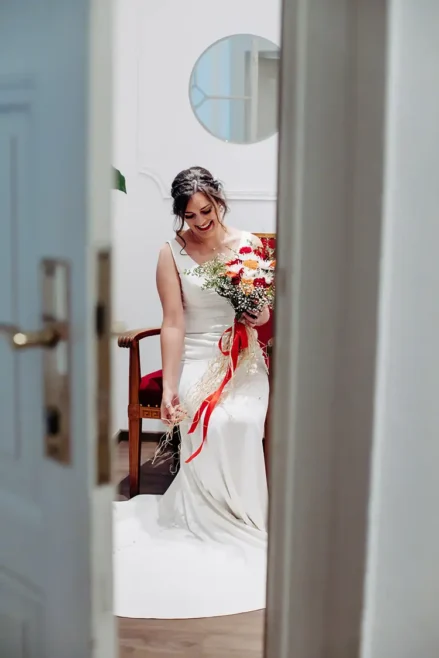 In einer Szene einer Hochzeit auf Schloss Albrechtsberg hält eine Braut in einem wallenden weißen Kleid einen Strauß roter und weißer Blumen mit Bändern in den Händen. Sie sitzt auf einem Stuhl in Dresden und blickt lächelnd nach unten. Die Szene wird von einem Türrahmen eingerahmt, an der Wand hinter ihr ist ein runder Spiegel zu sehen.