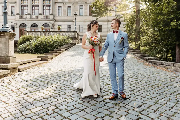 Eine Braut im weißen Kleid und ein Bräutigam im hellgrauen Anzug gehen Hand in Hand über einen Kopfsteinpflasterweg am Schloss Albrechtsberg, mit dem historischen Gebäude im Hintergrund. Sie lächeln sich an, im Sonnenlicht getaucht und umgeben von Dresdens üppigem Grün.