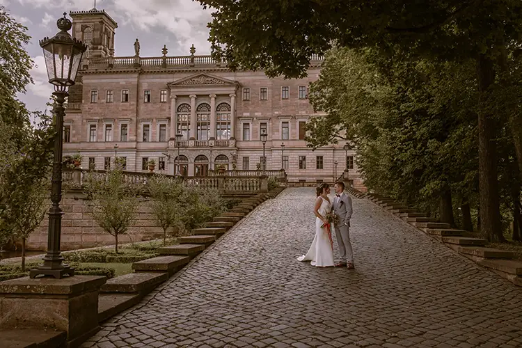 Braut und Bräutigam stehen auf einem Kopfsteinpflasterweg, der zum majestätischen Schloss Albrechtsberg führt. Das üppige Grün und der wolkenverhangene Himmel Dresdens schaffen eine romantische und zeitlose HOCHZEIT-Atmosphäre.