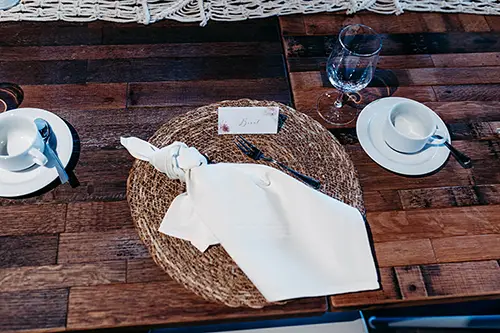 Ein rustikaler Holztisch ist mit zwei weißen Tassen auf Untertassen und jeweils einem Löffel gedeckt. Auf einem gewebten Tischset liegen eine ordentlich gefaltete Serviette und eine Namenskarte – perfekt für die Hochzeit im Berghotel Wilder Mann. Ein Weinglas daneben vervollständigt die elegante Tischdekoration.
