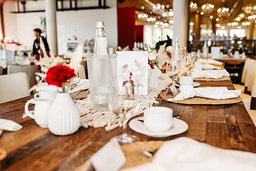 Für eine formelle Veranstaltung im Berghotel Wilder Mann ist ein wunderschön gedeckter Holztisch vorbereitet. Weißes Geschirr, Gläser, rote Rosen in Vasen und ein Tischläufer aus Spitze zieren den Tisch. Warme Beleuchtung und verschwommene Details im Interieur schaffen ein elegantes Ambiente.