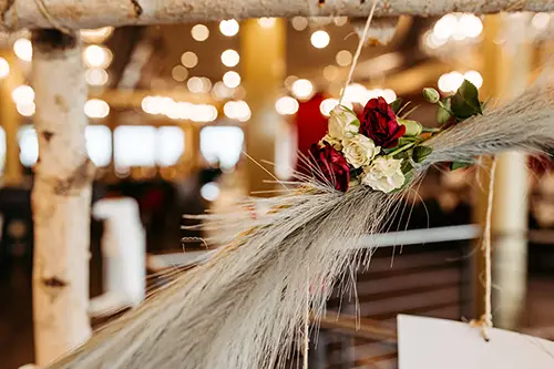 Eine Nahaufnahme der rustikalen Hochzeitsdekoration im Berghotel Wilder Mann zeigt einen Strauß roter und weißer Rosen, die mit beigen Weizenhalmen verflochten sind. Der Hintergrund ist sanft verschwommen und offenbart warmes Licht und eine gemütliche Innenatmosphäre.