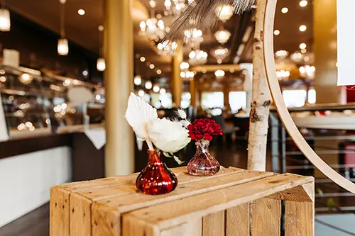 Kleine rote Vasen mit weißen und roten Blumen auf einer Holzkiste in einem warm beleuchteten Café versprühen den Charme einer Hochzeit im Berghotel Wilder Mann. Kronleuchter hängen von der Decke und schaffen eine einladende und gemütliche Atmosphäre.