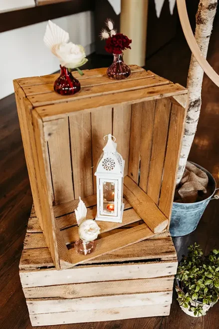 Holzkisten sind mit Dekorationen für eine Hochzeit im Berghotel Wilder Mann gestapelt. Auf dem Holzboden stehen eine weiße Laterne mit einer Kerze, kleine Vasen mit weißen und dunkelroten Blumen und eine kleine Pflanze in einem Blecheimer.