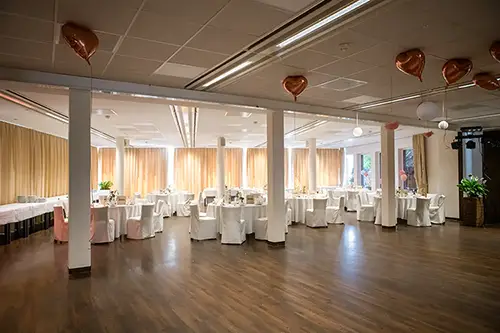 Ein großzügiger Bankettsaal mit Holzboden im Berghotel Wilder Mann ist für eine Veranstaltung bereit. Die Tische sind elegant mit weißen Tischdecken gedeckt, die Stühle mit passenden Hussen. Goldene herzförmige Ballons hängen von der Decke, und sanfte Beleuchtung schafft eine warme Atmosphäre.