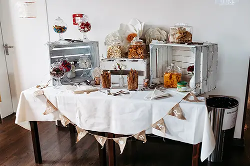 Eine dekorative Candybar auf der Hochzeit im Berghotel Wilder Mann mit verschiedenen Gläsern voller Süßigkeiten und Snacks. Holzkisten enthalten Gläser mit bunten Süßigkeiten und Leckereien. Ein Wimpel mit der Aufschrift „CANDY BAR“ schmückt elegant die Tischfront.