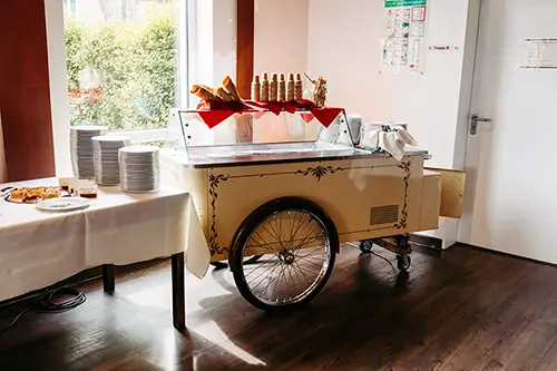Der Eiswagen steht drinnen neben einem mit Tellern und Gebäck beladenen Tisch – perfekt für eine Hochzeit im Berghotel Wilder Mann. Mit zwei Rädern und einer glänzenden Glasvitrine präsentiert er Eiswaffeln. Sonnenlicht fällt durch die Fenster und taucht die Szene in ein strahlendes Licht.