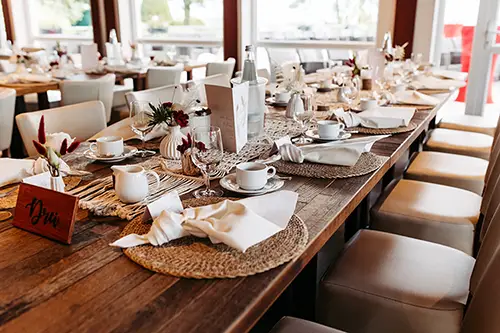Ein wunderschön gedeckter Esstisch mit elegantem weißen Geschirr, Gläsern und Blumenschmuck spiegelt die Essenz einer Hochzeit im Berghotel Wilder Mann wider. Sanftes Tageslicht strömt durch große Fenster und schafft eine warme und einladende Atmosphäre für Ihre besondere Feier.