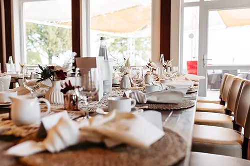 Ein wunderschön gedeckter Esstisch mit elegantem weißen Geschirr, Gläsern und Blumenschmuck spiegelt die Essenz einer Hochzeit im Berghotel Wilder Mann wider. Sanftes Tageslicht strömt durch große Fenster und schafft eine warme und einladende Atmosphäre für Ihre besondere Feier.