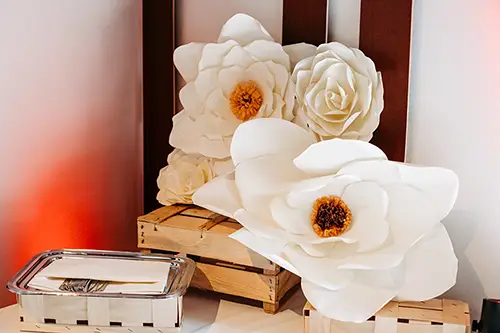 Große, weiße Papierblumen mit gelber Mitte sind elegant auf Holzkisten vor einer hellen Wand der Hochzeit im Berghotel Wilder Mann arrangiert. Links daneben steht ein silbernes Tablett mit einem kleinen Teller und Besteck, was dem Ambiente einen Hauch von Eleganz verleiht.