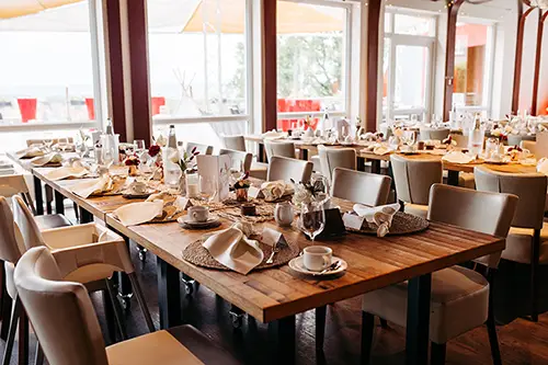 Der Speisesaal des Restaurants, ideal für eine Hochzeit im Berghotel Wilder Mann, besticht durch elegant gedeckte Holztische mit weißen Servietten und Gläsern. Durch große Fenster fällt Sonnenlicht auf die weiß gepolsterten Stühle und schafft eine einladende Atmosphäre, ideal für jede Feier.