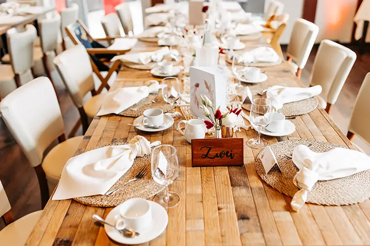 Ein elegant gedeckter Esstisch aus Holz für eine Hochzeit im Berghotel Wilder Mann ist mit weißen Servietten, Tassen und Weingläsern ausgestattet. Gedeckt werden gewebte Tischsets und ein Blumengesteck. Auf einer hölzernen Namenskarte steht „Love“, während cremefarbene Stühle den Tisch umgeben.