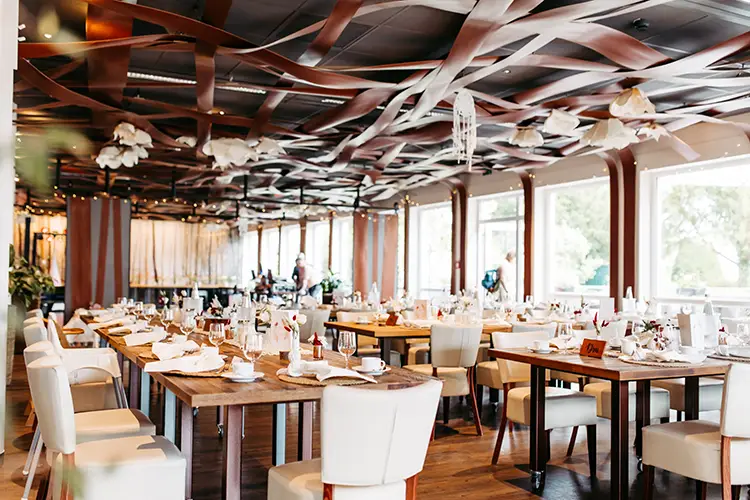 Elegantes Restaurant-Interieur mit gedeckten Holztischen, weißen Stühlen und kunstvoller Deckendekoration. Große Fenster bieten einen Blick ins Grüne und schaffen eine einladende und luftige Atmosphäre. Perfekt für eine Hochzeit im Berghotel Wilder Mann, aufgenommen von der Hochzeitsfotografin Dresden Melanie Kunert Fotografie.