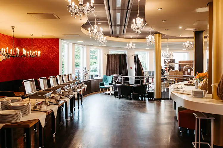 Ein geräumiger, eleganter Raum im Berghotel Wilder Mann, perfekt für eine Hochzeit, mit Buffet-Servierstationen, Kronleuchtern und großen Fenstern. Die Tische sind mit Tellern gedeckt, umgeben von roten, strukturierten Wänden und einem Mix aus modernen und klassischen Designelementen.