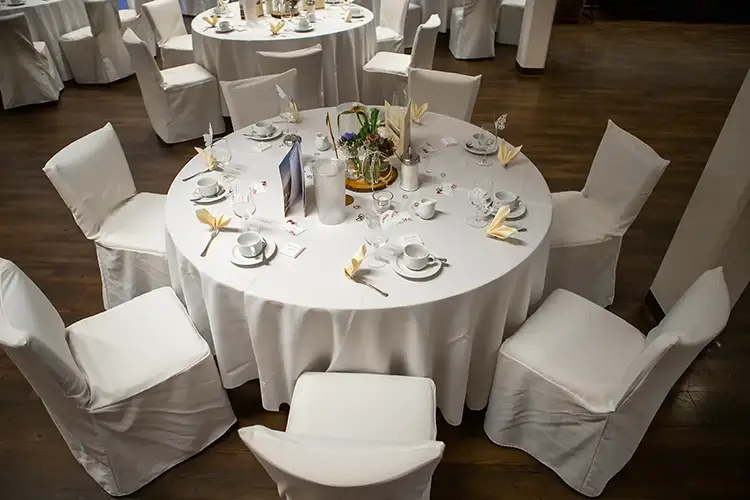 Ein runder Tisch bei der Hochzeit im Berghotel Wilder Mann, mit einer weißen Tischdecke bedeckt, ist elegant für eine Veranstaltung gedeckt. Acht Stühle mit passenden Hussen umgeben ihn. Teller, Tassen und Servietten sind ordentlich neben einem Blumengesteck und dekorativen Details von Melanie Kunert Fotografie arrangiert.