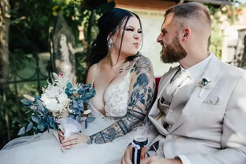 Braut und Bräutigam sitzen bei ihrer Hochzeit in Tharandt Seite an Seite und schneiden sich verspielt Grimassen. Die Braut, mit dunklem Haar und Tattoos, hält einen weißen Blumenstrauß in der Hand, während der Bräutigam im hellen Anzug einen Bart trägt. Beide wirken glücklich und entspannt in dieser bezaubernden Umgebung im Freien.