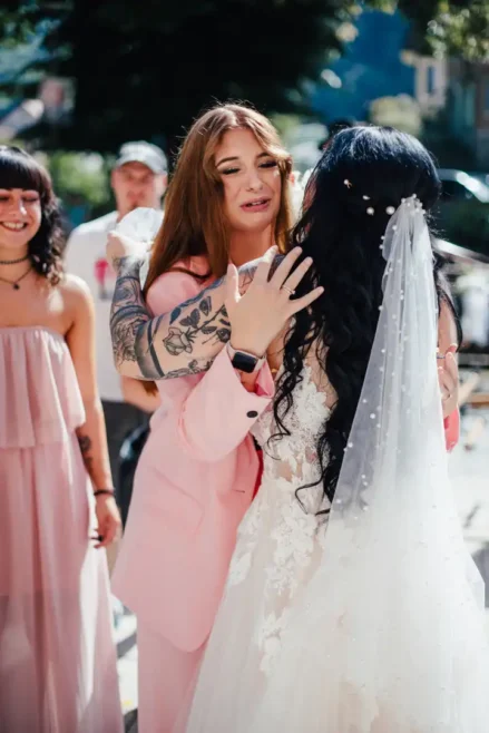 In einer sonnigen Außenkulisse fängt Melanie Kunert Fotografie eine freudige Umarmung bei einer Hochzeit in Tharandt ein. Eine Person im rosa Anzug, mit Tattoos und langen Haaren, umarmt eine andere in einem perlenbesetzten Brautkleid, während eine andere in einem passenden rosa Kleid strahlt.
