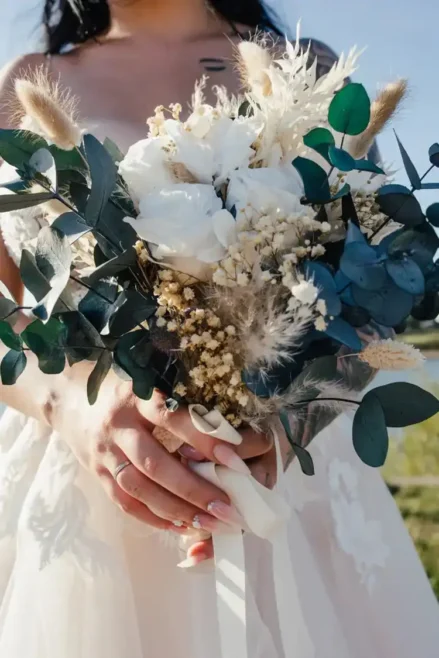 Die Braut steht anmutig da und hält einen Strauß weißer Blumen, Eukalyptusblätter und getrocknetes Laub in den Händen. Sie trägt ein weißes Kleid mit Spitzendetails, das ihre elegante Maniküre und Ringe vor dem Hintergrund eines strahlend blauen Himmels perfekt ergänzt – eine Bilderbuchhochzeit in Tharandt.