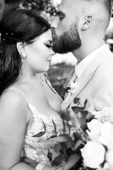 Ein Schwarz-Weiß-Foto zeigt eine heitere Hochzeit in Tharandt, während sich Braut und Bräutigam umarmen. Der Bräutigam im Anzug küsst die Braut sanft auf die Stirn, während sie die Augen schließt und leicht lächelt. In ihrem Spitzenkleid hält sie einen Blumenstrauß in der Hand, der ihre gemeinsame Zufriedenheit widerspiegelt.