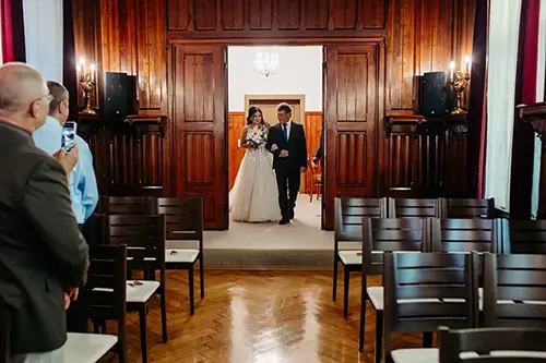 Eine Braut im weißen Kleid schreitet mit einem Mann im Anzug, vermutlich ihrem Vater, in einem holzgetäfelten Raum zum Altar. Gäste stehen zu beiden Seiten, einige halten Momente mit ihren Handys fest. Die Kulisse ist perfekt für eine Hochzeit in Tharandt – Melanie Kunert Fotografie fängt jedes Lächeln ein.