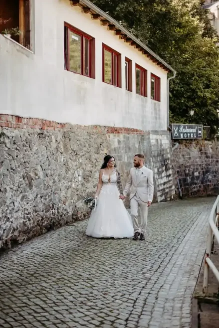 Eine Braut im weißen Kleid und ein Bräutigam im hellen Anzug schlendern Hand in Hand einen gepflasterten Weg entlang einer Steinmauer. Die Szene, aufgenommen von Melanie Kunert Fotografie, zeigt links ein Gebäude mit rot gerahmten Fenstern und im Hintergrund Bäume – eine unvergessliche Hochzeit in Tharandt.