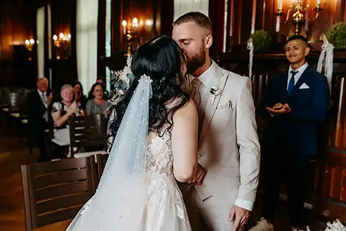 Bei einer Hochzeit in Tharandt, festgehalten von Melanie Kunert Fotografie, küssen sich Braut und Bräutigam in einem warm erleuchteten Raum zärtlich. Die Braut trägt ein weißes Kleid mit Schleier, der Bräutigam einen hellen Anzug. Die Gäste beobachten von ihren Plätzen aus, während der Trauzeuge daneben steht und applaudiert.