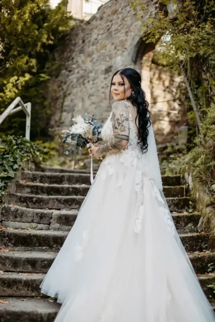 Die Braut im weißen Brautkleid, aufgenommen von Melanie Kunert Fotografie während einer Hochzeit in Tharandt, steht anmutig auf Steinstufen im Freien. Ihr langes dunkles Haar fällt über sichtbar tätowierte Arme. Eine alte Steinmauer und üppiges Grün runden die bezaubernde Szene ab.