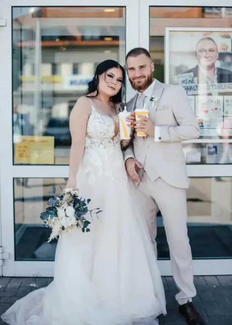Braut und Bräutigam stehen vor einem Schaufenster i, lächeln sich an und halten Erfrischungsgetränke in den Händen. Die Braut trägt ein Brautkleid aus floraler Spitze und hält einen Blumenstrauß in der Hand, während der Bräutigam einen hellen Anzug mit einer Blumenansteckblume trägt. Ein bezaubernder Hochzeitsmoment, perfekt eingefangen.